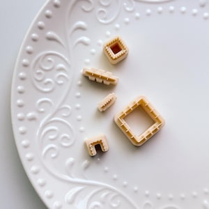 May include: A collection of beige plastic cookie cutters in various geometric shapes, including squares and rectangles, are arranged on a white, ornate plate. The cutters have a textured edge, and the plate has a decorative floral pattern.