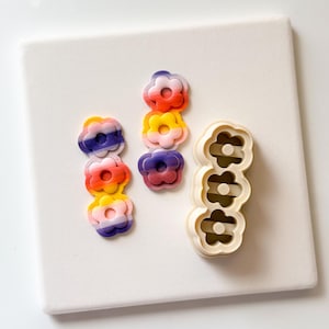 Puede incluir: Una colección de pendientes de arcilla polimérica con forma de flor y un cortador a juego. Los pendientes son en tonos rojo, naranja, amarillo, morado y azul. El cortador es de color beige.