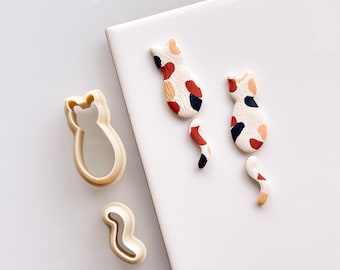 May include: Two clay cat figurines with a white base and red, orange, and black spots. The cats are next to two plastic cat cookie cutters, one of a cat and one of a tail.