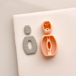 Könnte beinhalten: Zweiteilige ohrringförmige Polymer Clay Ausstecher. Ein Set ist grau mit einem floralen Design, das andere ist ein pfirsichfarbener Kunststoffausstecher. Die Ausstecher sind für die Herstellung von Ohrringen bestimmt.