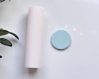 以下が含まれることがあります： テクスチャ加工された表面を持つ淡いピンク色の円筒形の物体と、模様のある表面を持つ小さく丸い水色の物体。白い表面に置かれ、背景には緑の葉があります。
