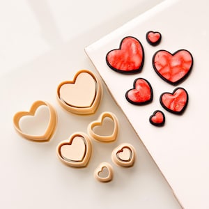 May include: A set of heart-shaped cookie cutters in various sizes, ranging from small to large. The cutters are made of a beige plastic material. There are also several red heart-shaped cookies on a white surface.