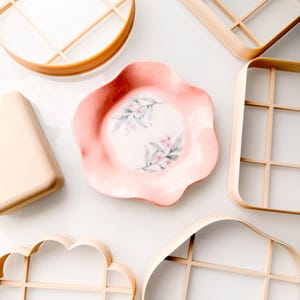 May include: A central pink flower-shaped ceramic dish with a delicate floral pattern surrounded by several beige plastic geometric and organic-shaped cookie cutters featuring internal grid structures for creating precise shapes.