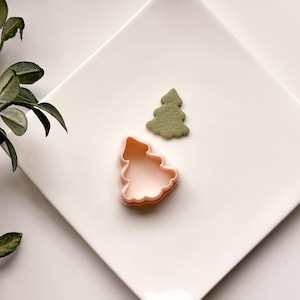 Puede incluir: Dos cortadores de galletas de árbol de Navidad de plástico en un plato blanco. El cortador más grande es rosa y el más pequeño es verde.