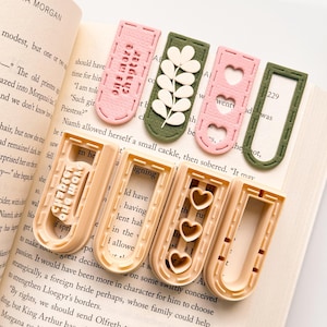 以下が含まれることがあります： 様々なデザインの手作りブックマークのコレクション。ピンクとグリーンの長方形で、ステッチされたエッジがあります。ハートの切り抜き、花のアクセント、または「one more chapter」のテキストが特徴です。その他はベージュ色で、U字型で、ハートの切り抜きがあります。