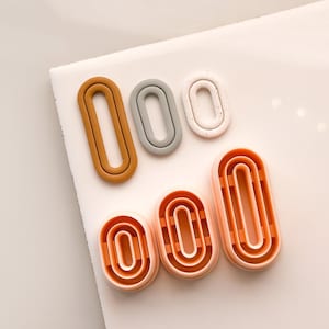 May include: A collection of oval-shaped cookie cutters in various sizes and colors, including brown, gray, white, and shades of orange. The cutters are arranged on a white surface.