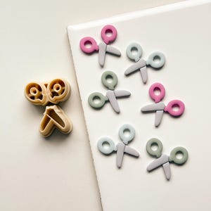 May include: A collection of colorful scissor-shaped cookie cutters and a matching cookie cutter. The scissors are in shades of pink, green, and blue, with gray blades. The cookie cutter is tan.