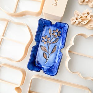 May include: A rectangular blue ceramic tray with a debossed floral design featuring two flowers and leaves, surrounded by several beige plastic cookie cutters of various shapes, including one labelled Lulu Cutter in embossed lettering.
