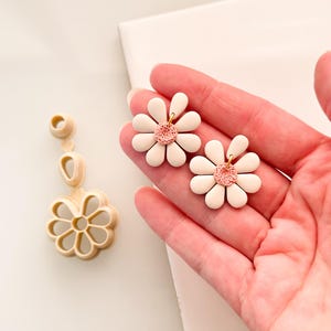 Könnte beinhalten: Ein Paar weiße Gänseblümchen-Ohrringe mit rosafarbenen Zentren. Die Ohrringe sind aus Polymer Clay gefertigt und haben einen goldenen Haken.