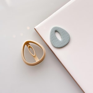 May include: A light brown polymer clay cutter and a light blue polymer clay earring with a textured pattern. The cutter is in the shape of an oval with a hole in the center. The earring is also oval-shaped.