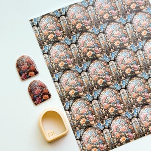 Puede incluir: Una hoja de papel con un patrón floral de vidrieras, dos calcomanías para uñas y un cortador de forma de uñas beige. Las calcomanías y el patrón presentan flores rosas, naranjas y azules. El cortador de forma de uñas tiene el número "601" impreso.