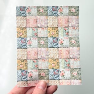 May include: A rectangular sheet featuring a patchwork design with floral and lace patterns. The squares are in pastel colors like pink, blue, and yellow, with a vintage aesthetic. The sheet is held by a hand.