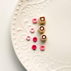 May include: A white, ornate plate holds an assortment of small, heart-shaped and geometric-shaped candies. The candies are in shades of pink and brown. Some candies have heart-shaped cutouts. The plate has a decorative, embossed design.