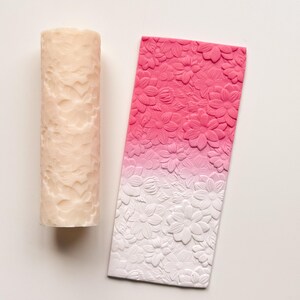 May include: A cylindrical candle and a rectangular decorative piece, both featuring a floral pattern. The candle is a light beige color, while the rectangular piece has a gradient of pink to white, with the flowers in relief.