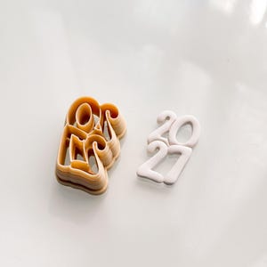 May include: A light tan plastic cookie cutter shaped like the number 2027, featuring a thick frame and open interior design, positioned next to a white fondant or dough cutout of the same number 2027 with rounded, smooth edges.