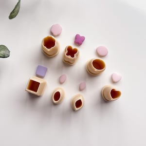 Puede incluir: Un conjunto de cortadores de galletas beige en varias formas, incluyendo círculos, cuadrados, corazones y una mariposa. Acompañando a los cortadores hay pequeñas formas rosas y moradas. Los artículos están dispuestos sobre una superficie blanca.