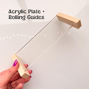 May include: A clear acrylic plate with two beige rolling guides attached to the sides. The plate is held in a hand.