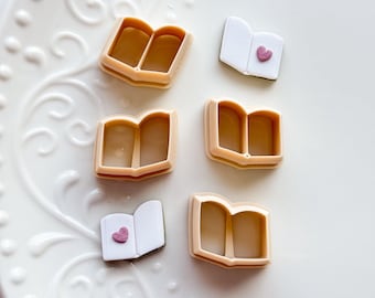 May include: A collection of tan plastic book-shaped cookie cutters and open book-shaped fondant decorations with pink heart accents, arranged on a white, ornate plate. The cutters are open, showing the interior shape.