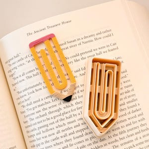 May include: Two yellow and pink pencil-shaped bookmarks with a textured surface. The bookmarks are placed on top of an open book with text about a castle and a great hall.