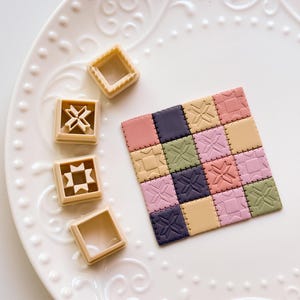 May include: A colourful patchwork quilt composed of small squares with embossed patterns. Next to it are three beige square cookie cutters with star and geometric designs. The items are on a white plate with a decorative edge.