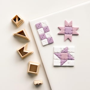 May include: An overhead shot displays quilting tools and finished quilt block designs. The tools are tan plastic, and the blocks feature white, lavender, and pink colors. The blocks are in various shapes, including a star and a square.