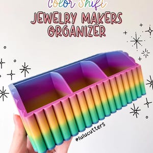 May include: A rainbow color-shifting jewelry organizer with three compartments. The organizer is made of plastic and has a ribbed texture. The text "color shift JEWELRY MAKERS ORGANIZER" is written in pink, yellow, green, and blue letters on a white background.