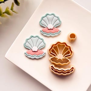 Puede incluir: Un juego de dos cortadores de galletas con forma de concha de mar con una perla en su interior. Los cortadores son de plástico y de color marrón claro. Hay dos galletas terminadas en un plato blanco, una es azul claro y la otra rosa.