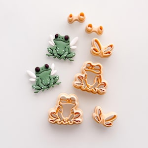 May include: An assortment of frog-themed items. There are three green frog figurines with white wings and white spots. Also included are several tan-colored frog and butterfly-shaped cookie cutters. The items are arranged on a white surface.