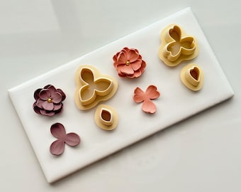 May include: A set of flower-shaped cookie cutters and fondant flowers in shades of pink and brown. The cutters are made of plastic and are arranged on a white tile surface.