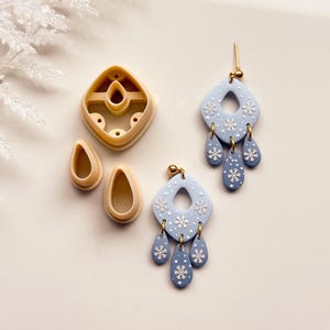 May include: A pair of light blue dangle earrings with white snowflake details and gold-colored hardware. The earrings are diamond-shaped with three teardrop-shaped dangles. Also shown are polymer clay cutters in beige, including a diamond shape and two teardrop shapes.