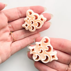 May include: Two pale peach-colored clay cutters featuring a repeating celestial pattern of crescent moons and four-pointed stars. The cutters have a raised, rounded edge design and are shaped to create multiple small shapes in a single press.