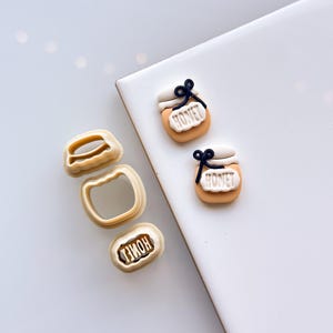 May include: A collection of biscuit-coloured cookie cutters and honey jar decorations. The cutters are shaped like a jar and the word "HONEY". The honey jars feature a white lid, a black bow, and the word "HONEY" on a label.