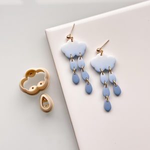 Puede incluir: Un par de pendientes de nube azul y blanco degradado con detalles dorados y un juego de cortadores de arcilla a juego para hacer los pendientes.
