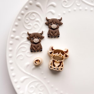 May include: A selection of Highland cow-themed items, including three brown cow-shaped decorations and a cow-shaped cookie cutter. The decorations have a smiling face and are sitting down. The cookie cutter is beige and brown.
