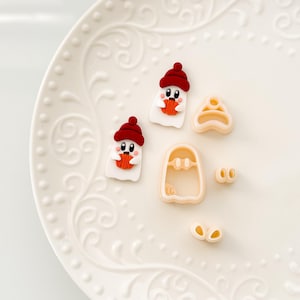 May include: A white ceramic plate with a decorative pattern displays Halloween-themed polymer clay figures. Two ghost figures wear red hats and hold orange pumpkins. There are also ghost-shaped cookie cutters and eye-shaped cutters.