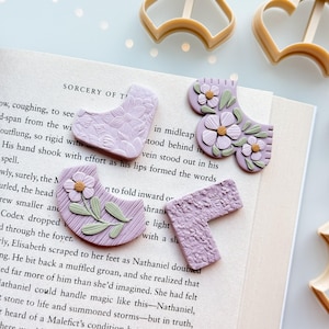 May include: Polymer clay cutters and floral-decorated clay pieces in lavender, green, and white, arranged on an open book. The cutters are tan-coloured, and the book's pages are visible.