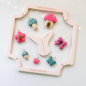May include: A collection of colorful polymer clay charms, including mushrooms, flowers, butterflies, and a bow, displayed on a pink frame. The frame is labeled "Lulu Cutters" and "Bead Guide".