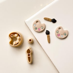 May include: A collection of beige-colored art-themed cookie cutters and decorative elements. The set includes palette shapes with pink, blue, and green circles, paintbrushes, and a round object. The items are arranged on a white surface.