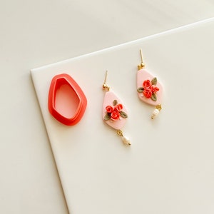 May include: A pair of pink and gold earrings with a floral design. The earrings are made of polymer clay and feature a cluster of red roses with green leaves. Each earring has a small pearl dangling from the bottom.