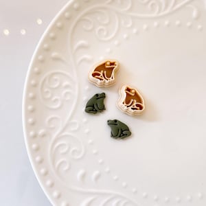 May include: Four frog-shaped cookie cutters in shades of green and tan are arranged on a white, decorative plate. Two cutters are green, and two are tan with a brown outline. The plate has a detailed, embossed floral design.