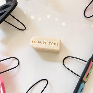 May include: A light beige LC Wide Tool is surrounded by several black and colored hair clips with metal arches. The text "LC WIDE TOOL" is embossed on the tool. The clips are in various colors, including pink, white, and black.