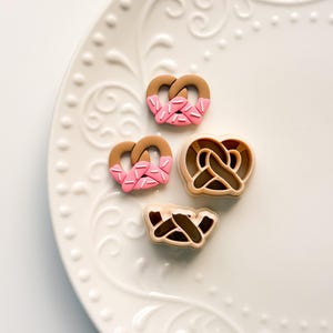 Puede incluir: Cuatro elementos en forma de pretzel sobre un plato blanco estampado. Dos son marrones con glaseado y chispas rosas, y dos son cortadores en forma de pretzel. El plato tiene un diseño decorativo en relieve.