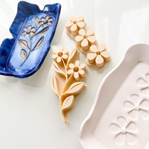 May include: A rectangular blue ceramic tray with a floral relief, a rectangular white ceramic tray with a debossed flower pattern, and two beige plastic flower-shaped cookie cutters with a stem and leaf design, arranged on a flat surface.