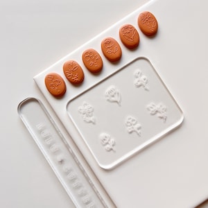 May include: A set of six floral polymer clay cutters with a clear acrylic sheet featuring the same floral designs. The cutters are orange and the sheet is clear. The sheet has the text "Lulu Cutters" printed on it.