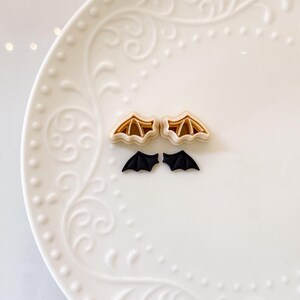 May include: Two sets of bat-shaped cookie cutters. The top set is tan with a brown interior, and the bottom set is black. The cutters are arranged on a white, patterned plate, perfect for Halloween baking.