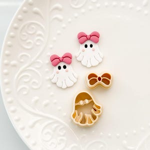 May include: A collection of Halloween-themed cookie cutters and decorations. Two white ghost-shaped decorations with pink bows and black dot eyes are displayed with a bow-shaped cutter and a ghost-shaped cutter. The items are on a white, patterned plate.