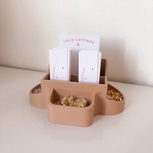 Könnte beinhalten: Ein hellbrauner, 3D-gedruckter Ohrring-Organizer mit drei Fächern. Das obere Fach enthält Visitenkarten mit dem aufgedruckten Text "LULU-CUTTERS". Die beiden unteren Fächer enthalten Ohrringverschlüsse.