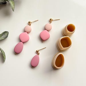 Puede incluir: Un conjunto de tres pendientes de arcilla rosa y rosa claro con postes dorados. Los pendientes se muestran sobre una superficie blanca con una pequeña caja marrón.