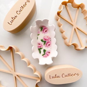 Peut inclure: Deux tampons ovales beiges avec le texte Lulu Cutters, deux emporte-pièces beiges à bords festonnés avec croisillons internes, et un plat ovale blanc central orné d'un motif floral de pivoines roses et de feuilles vertes sur fond blanc.