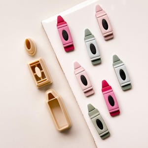 May include: A collection of colorful crayon-shaped objects in various shades of pink, green, and beige. The crayons have a black oval detail in the center. There are also beige crayon-shaped molds.
